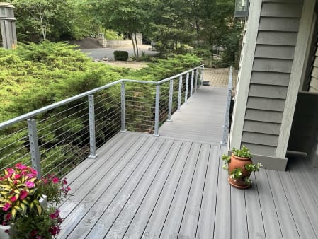 Legacy gray cable railing lines a composite walkway deck beside a shingled home and evergreen shrubs.