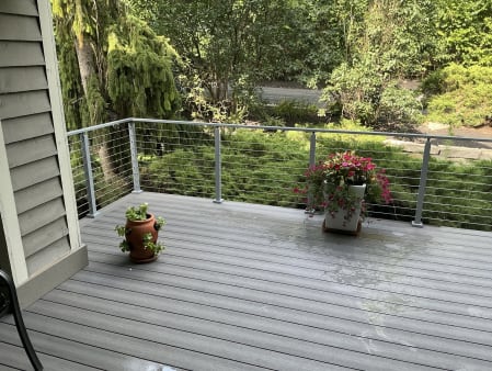 A gray composite deck with flower pots edged by gray cable railing against lush shrubs.