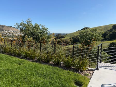 Black horizontal rod railing along a planted garden bed and lawn with green hills beyond.