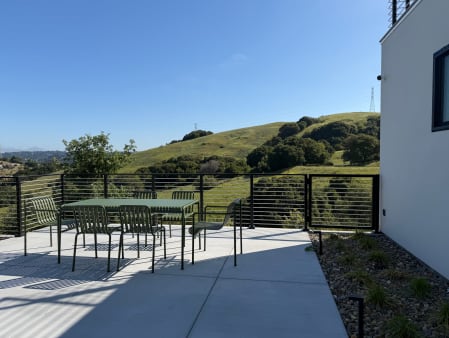 Modern home patio with outdoor dining set enclosed by black rod railing above golden-green hills.