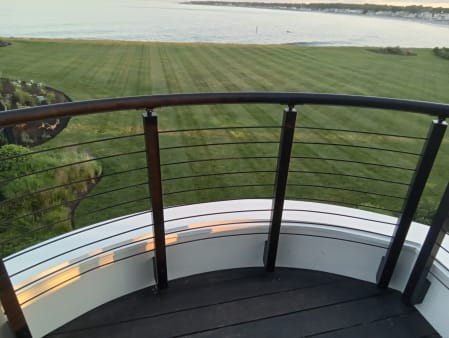 Curved black round handrail with cable infill wraps a turret balcony overlooking a lawn and the sea.