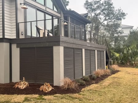 Black rod railing runs along a raised deck above louvered skirting on a coastal home.