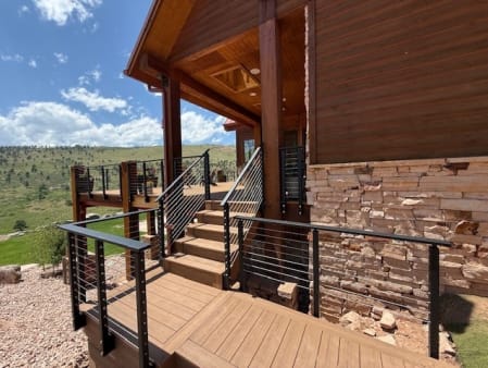 Black cable railing follows exterior deck stairs and landing beside a stone and wood mountain home.