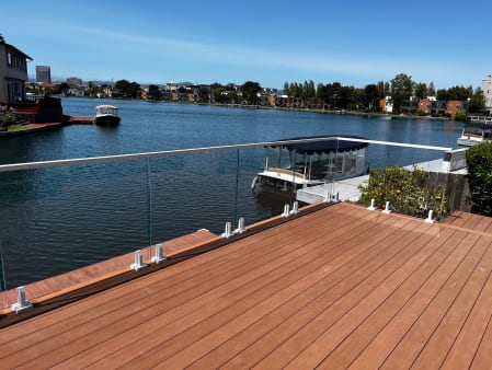 Frameless glass on white spigots with a white top cap edges a wood deck over a calm waterway.
