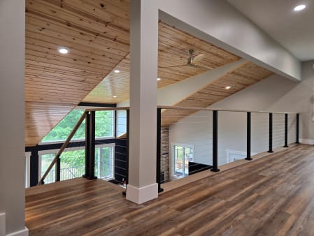 Black-post cable railing with a wood cap rail guards an open loft beneath a vaulted knotty-pine ceiling.