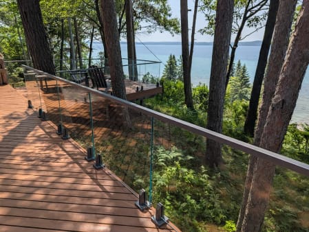 Glass panels on spigots with a top cap rail run along a wood deck high among pines above a lake.