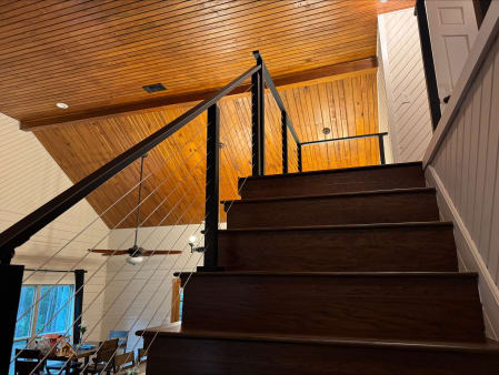 Black cable railing climbs a dark wood stair to a loft under a warm pine ceiling in a cabin.