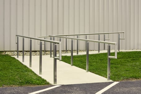 Brushed stainless post-mounted handrails lining a concrete accessibility ramp beside a metal building.