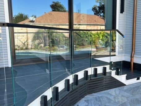Frameless glass on black side-mount talons curves along a pool deck beside a sliding glass wall