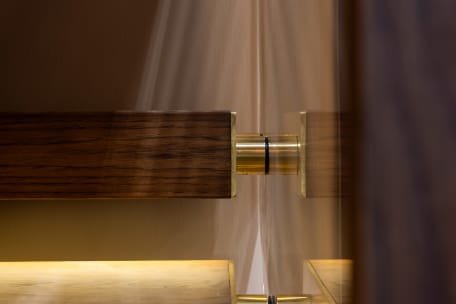 Close-up of a brass standoff pin securing glass beside a dark wood handrail with warm LED light below.