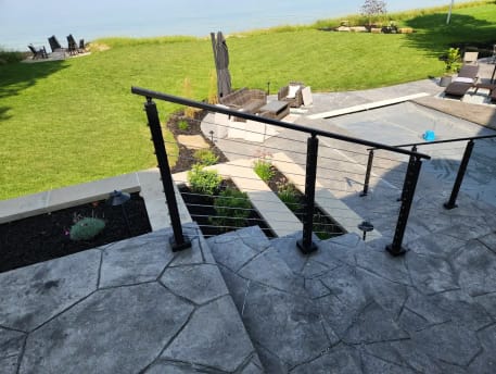 Black cable railing guards wide stamped-concrete steps descending to a lawn and lakefront patio.