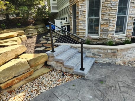 Short black cable railing flanks stone garden steps beside boulders and a stone-veneer wall.