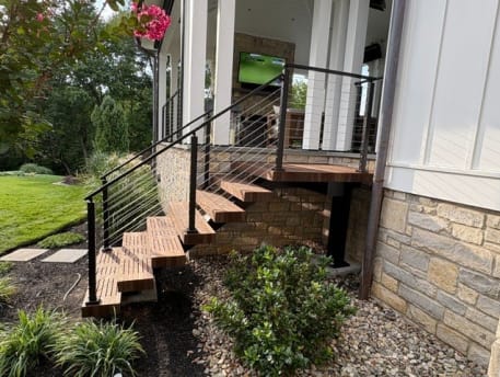 Thick hardwood treads climb to a covered porch with black-post cable railing beside stone veneer and lush plantings.