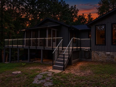 Dark-sided cabin with a wraparound deck and stair lined in black cable railing with lit handrail at sunset.
