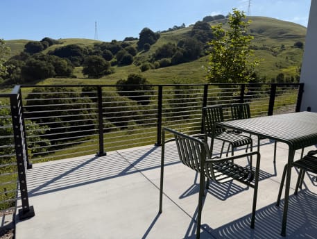 Concrete patio with green bistro chairs edged by black horizontal rod railing overlooking rolling green hills.