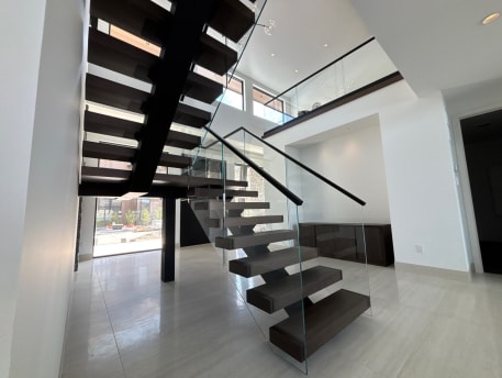 Underside view of dark wood floating treads on black mono stringers with frameless glass railing in a bright white hall.