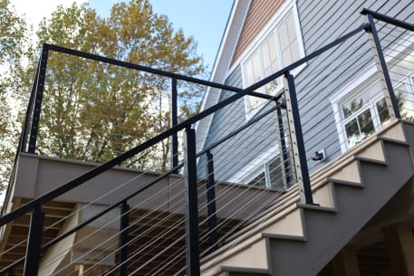 Black cable railing climbs an exterior deck stair beside a blue-sided home under autumn trees.