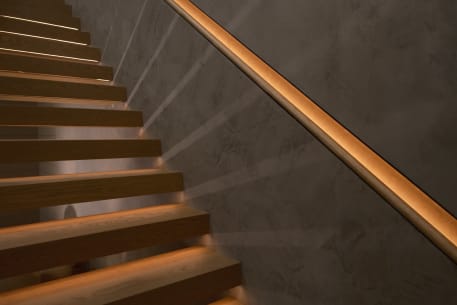 Wood handrail set into a lit wall reveal runs above oak floating treads against a plaster wall.