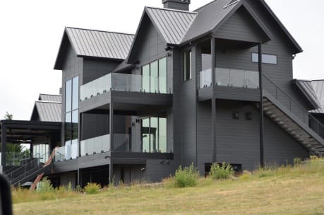 Frameless glass railings wrap stacked balconies on a dark modern farmhouse with metal roofs.