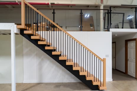 Side view of an oak-tread floating stair on a black stringer with black balusters and oak handrail along a white paneled wall.