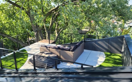 Black cable railing encloses a rooftop terrace with turf, pavers and an outdoor sectional among tree canopy.
