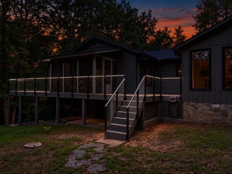 Dark-sided cabin with a wraparound deck and stair lined in black cable railing with lit handrail at sunset.