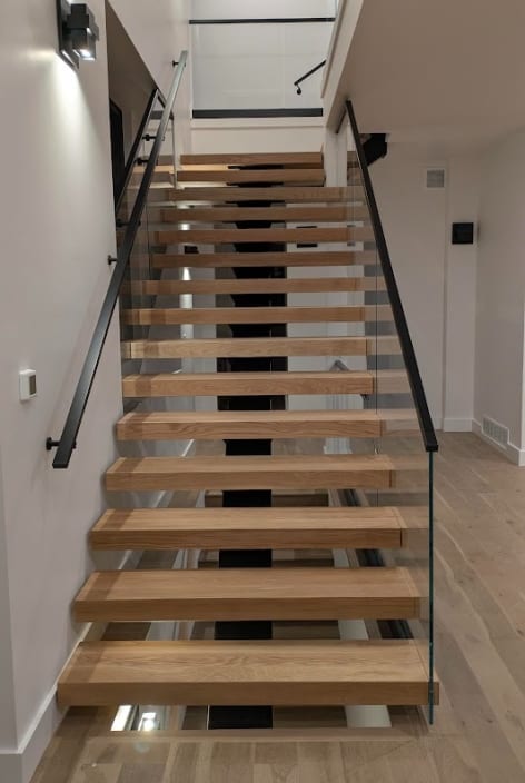 Straight run of white oak floating treads on a mono stringer with frameless glass on one side and a black wall-mounted handrail on the other.
