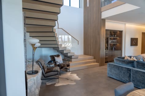 Light oak cantilevered treads rise from stacked plinth steps beside a glass railing in a modern living room.