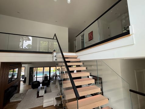 Oak floating treads with a black handrail and frameless glass rising to a glass-railed loft above a living room.