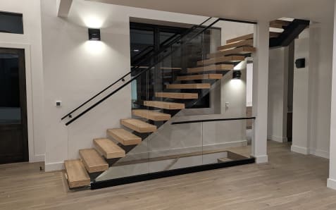 Oak floating treads on a black mono stringer with frameless glass and wall handrail beside a window in a bright great room.