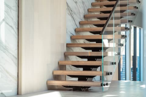 Oak treads on a black mono stringer with glass on black standoff pins beside a marble-clad wall.