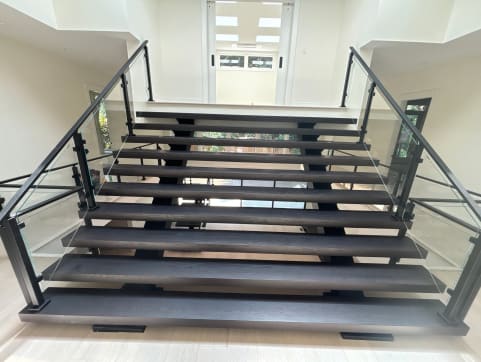 Dark wide floating treads on dual stringers with black-post glass railings rise toward a bright skylit hallway.