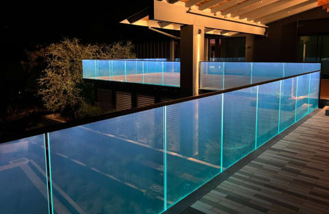 Glass base rail railing with an LED-lit top cap glows blue along a wood rooftop terrace at night.