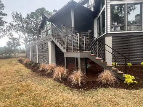 Horizontal rod railing follows an exterior stair and wraps a raised deck above landscaped beds.