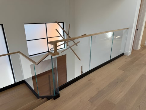 Frameless glass in a black floor channel with a light wood handrail around a stair opening with oak floors.