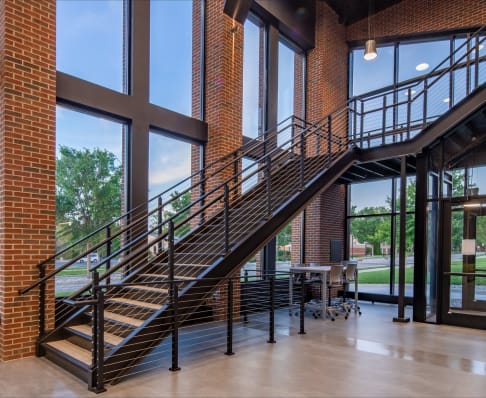 Black steel stair with cable railing and grab rail in a double-height brick-and-glass commercial lobby.