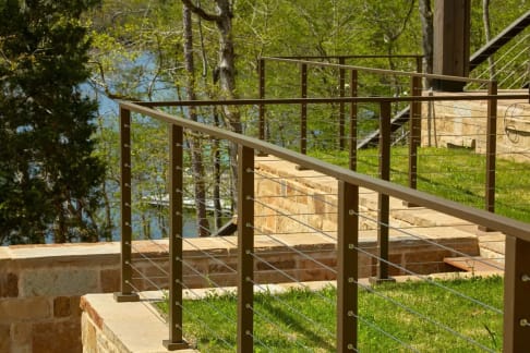 Bronze-finish posts and top rail with cable infill along a stone retaining wall above a lawn and lake.