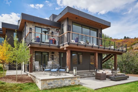 Cable railing with wood cap wraps the upper deck of a stone-and-timber modern home above a furnished patio.