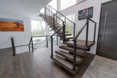 Stained oak floating treads with black cable railing turn at a landing beside tall windows.