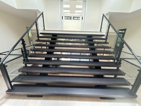 Dark wide floating treads on dual stringers with black-post glass railings rise toward a bright skylit hallway.