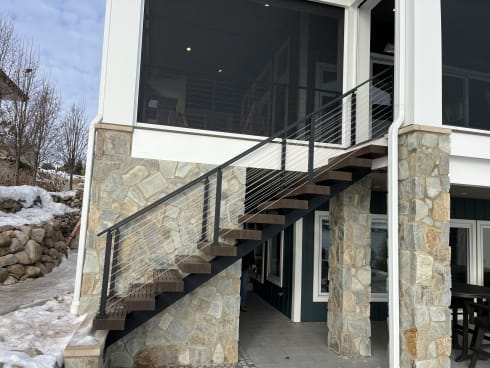 An exterior steel mono-stringer stair with dark treads and black cable railing climbs past stone columns to a screened porch.