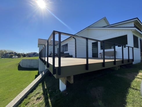 Black slim side-mount cable railing with double corner posts wraps a raised deck on a white farmhouse.
