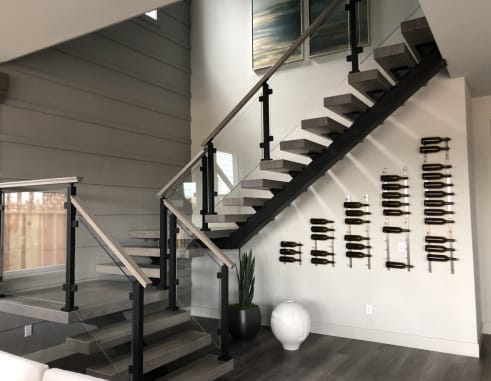 Black mono-stringer floating stair with gray treads and post-to-post glass railing beside a wine-bottle wall display.