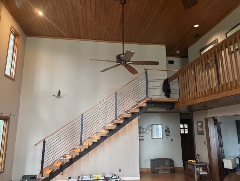 Pine floating treads on a black mono stringer with black cable railing and pine handrail rising to a loft under a tongue-and-groove ceiling