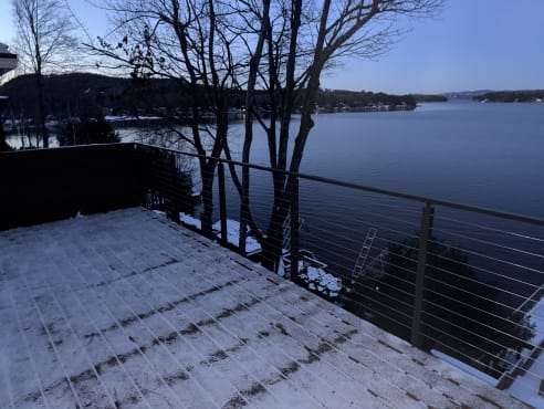 Snow-dusted deck with black cable railing overlooking a lake at dusk