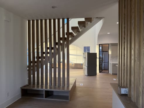 A floating oak stair with glass railing rises behind a vertical wood slat screen and bench in an open kitchen space.