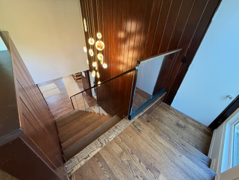 A frameless glass railing with a dark handrail guards the top of an oak stair beside dark wood paneling.