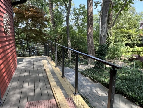 A gray deck beside a red house edged with black cable railing amid lush trees.
