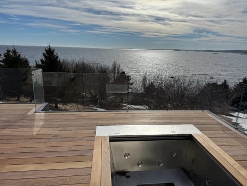 Frameless glass railing around a hardwood rooftop deck with hot tub overlooking the ocean at sunset.