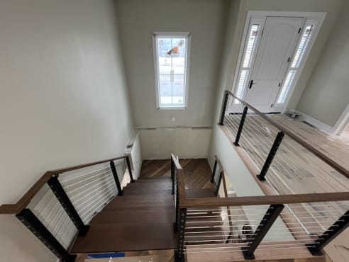 Black-post cable railing with a wood handrail surrounds an open wood stair and landing in a bright entry.